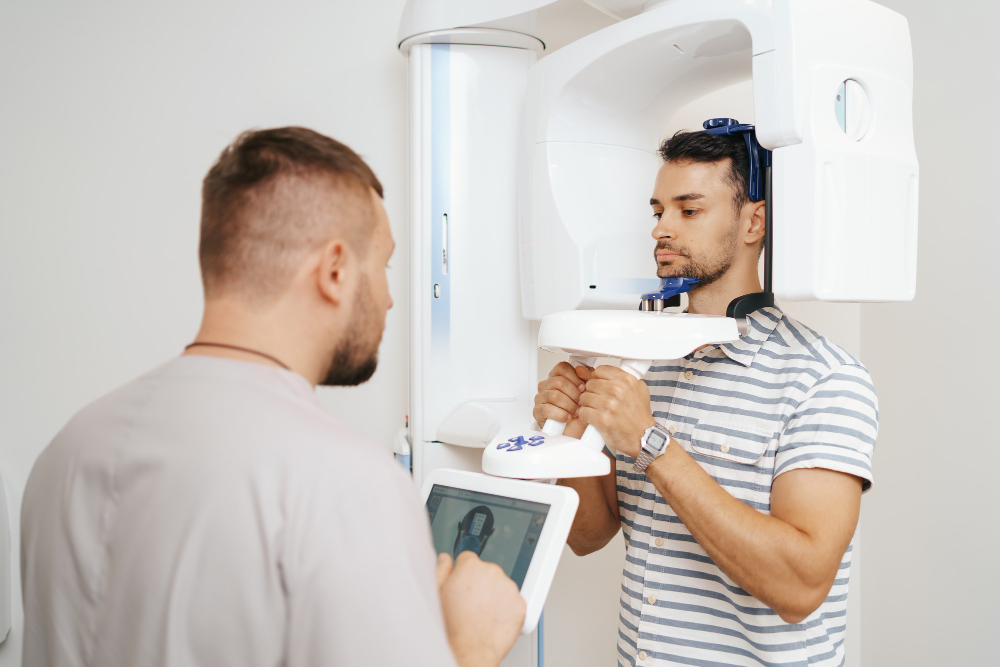 Man undergoing a dental X-ray scan while holding handles on the machine, with a technician operating the control panel.