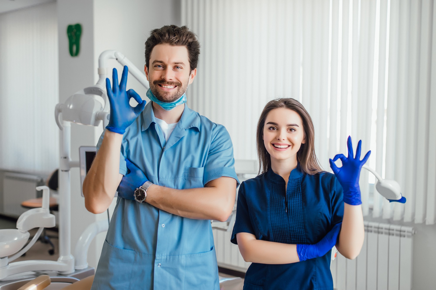 Male and female dental professionals smiling and wearing blue scrubs and gloves, both showing the OK hand gesture in a dental clinic.