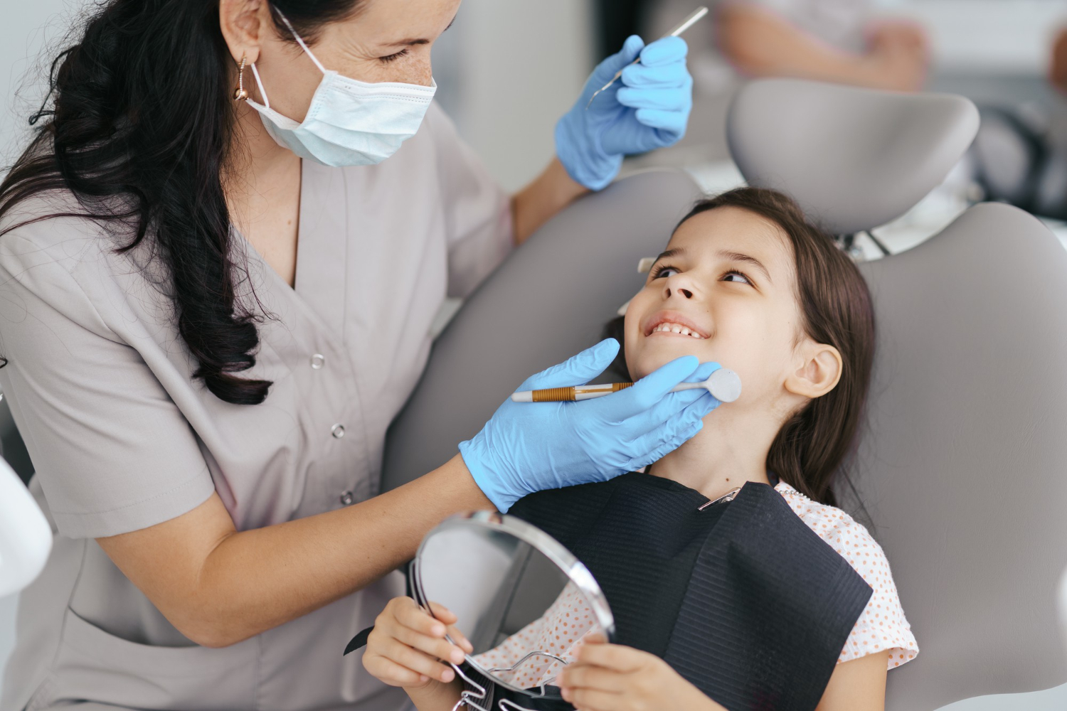 Dentist wearing mask and gloves examining smiling young girl sitting in dental chair holding a mirror.