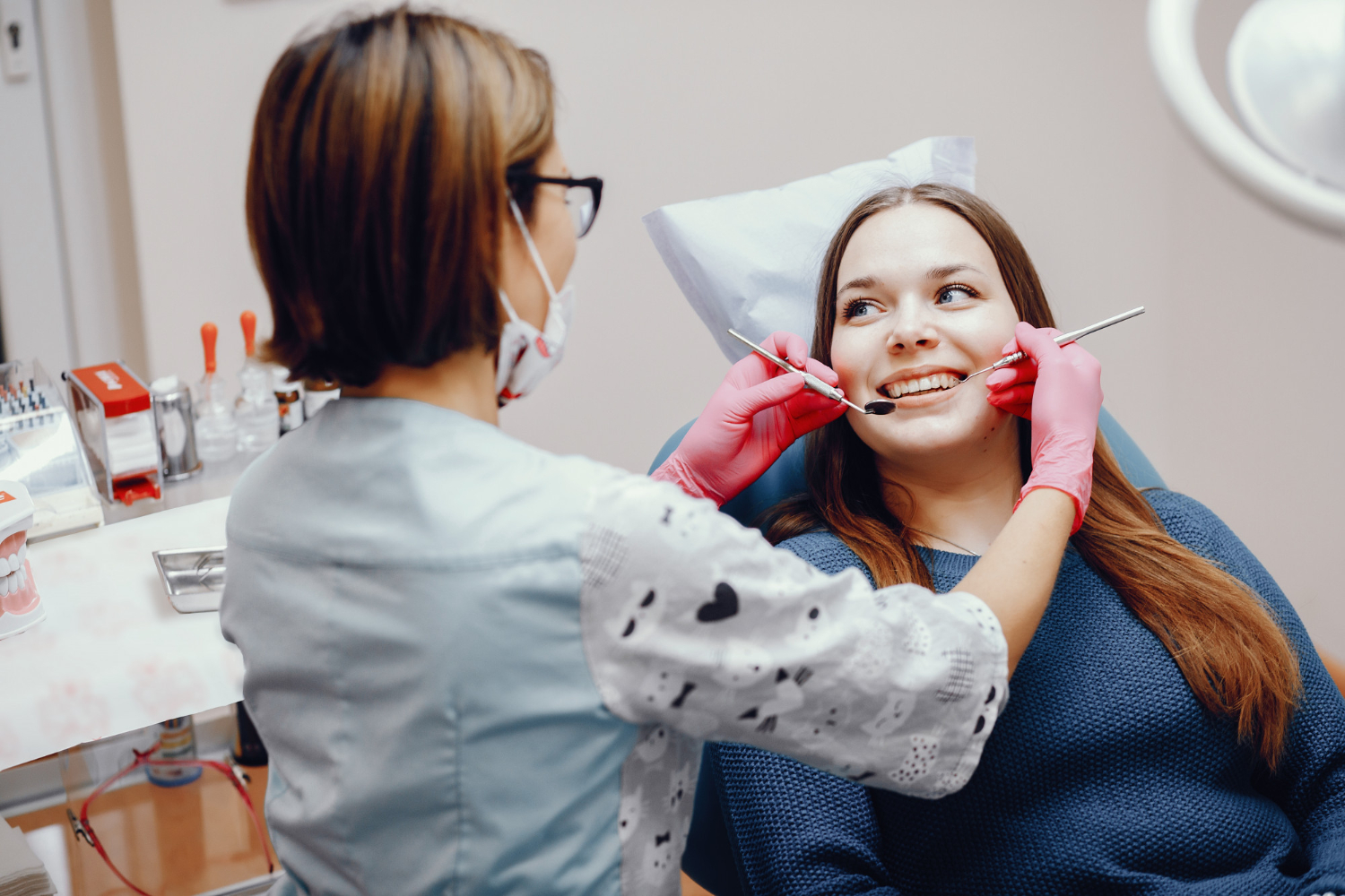 Dentist in pink gloves examining a smiling woman's teeth with dental instruments in a clinic.