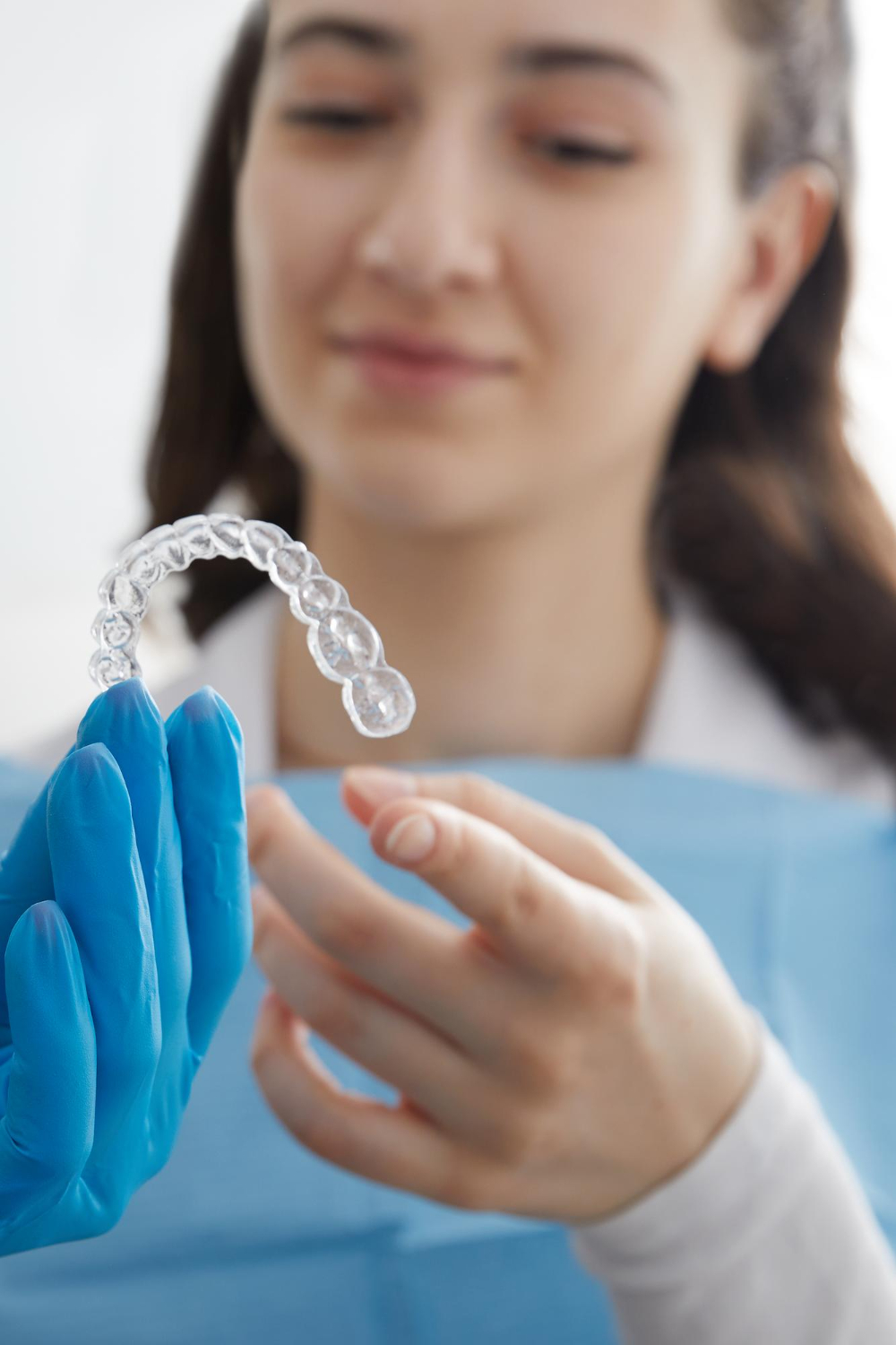 Close-up of a gloved hand holding a clear dental aligner with a blurred woman in a dental bib in the background.