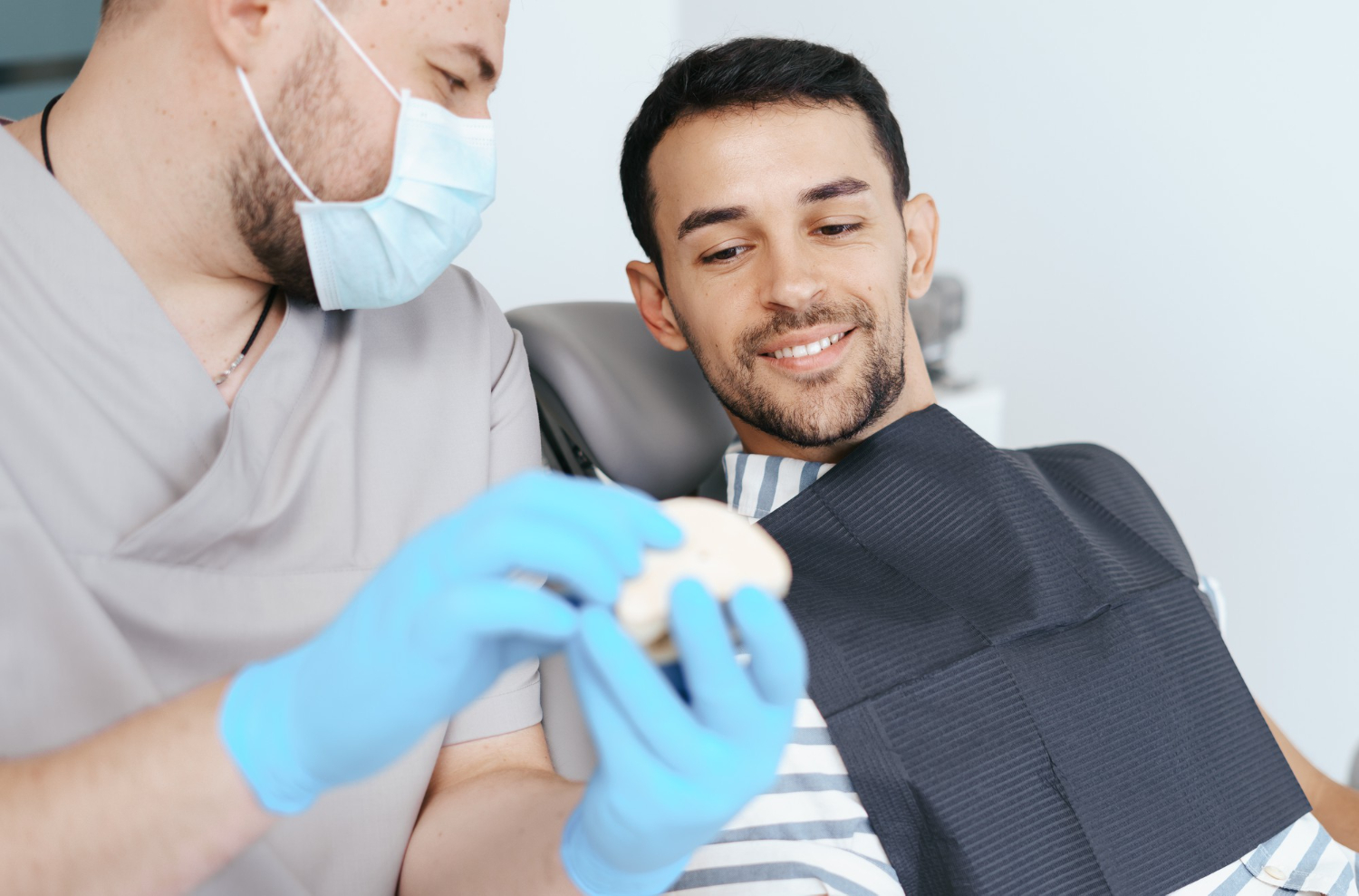 Dentist wearing a mask and gloves showing a dental model to a smiling male patient in a striped shirt with a dental bib.