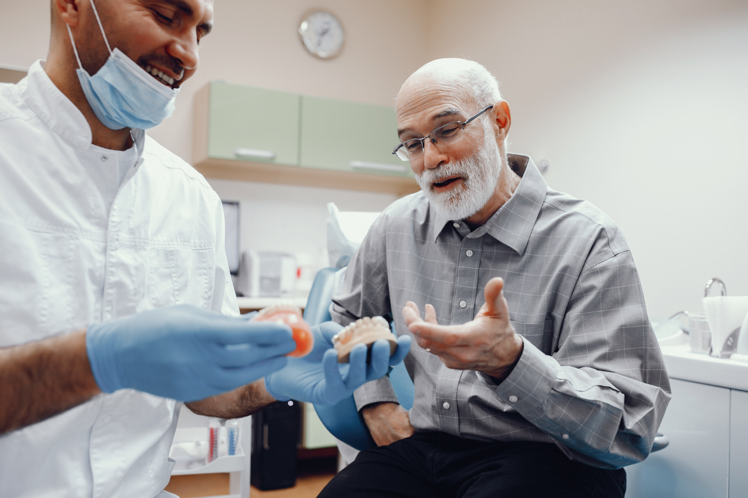 Dentist wearing blue gloves and mask holding a dental model and explaining to an elderly man with glasses in a dental office.