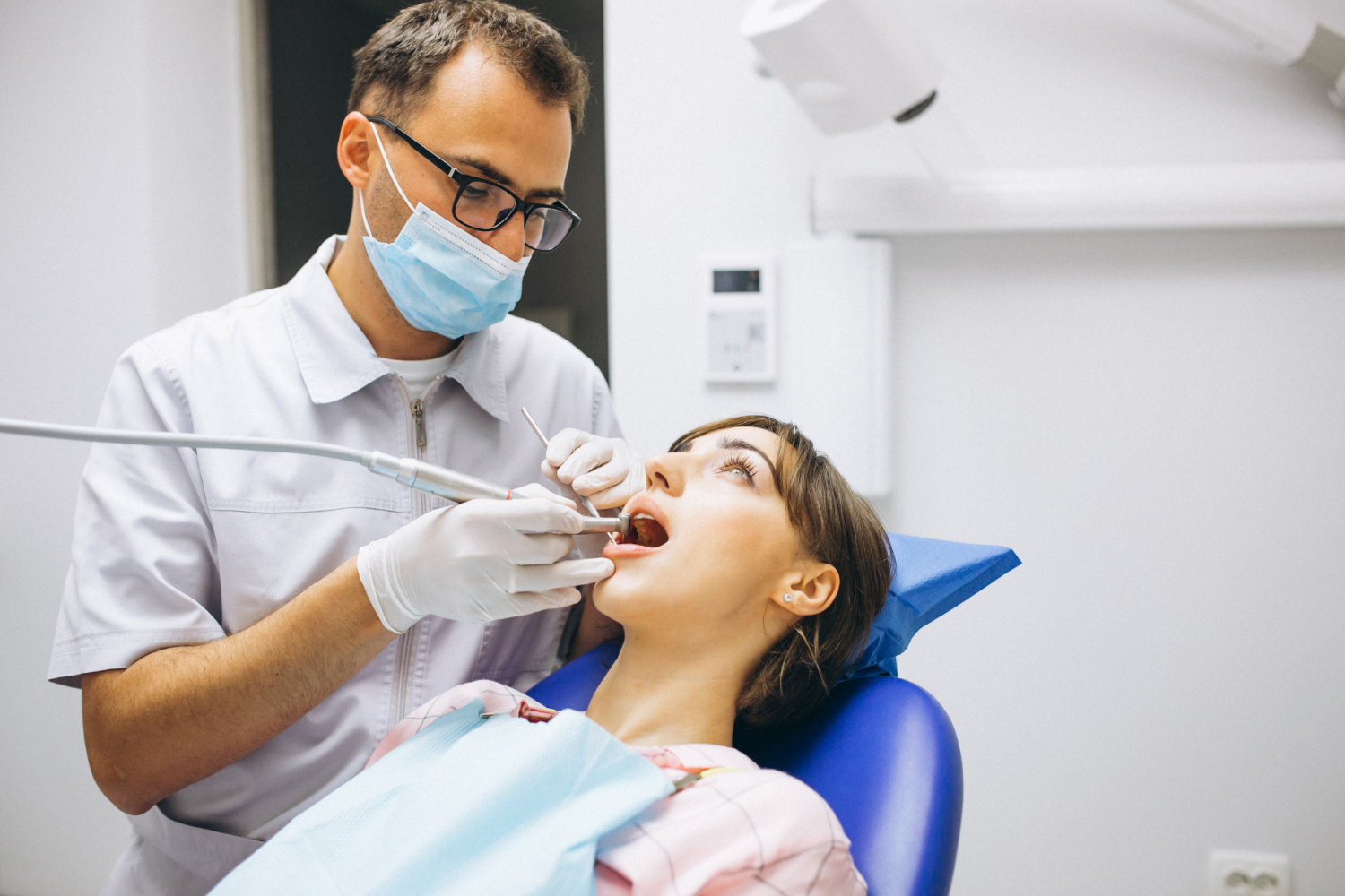 Dentist wearing glasses and a mask treating a female patient reclining in a dental chair.