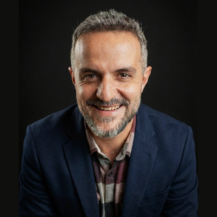 Smiling man with short hair and beard wearing a plaid shirt in an indoor setting.