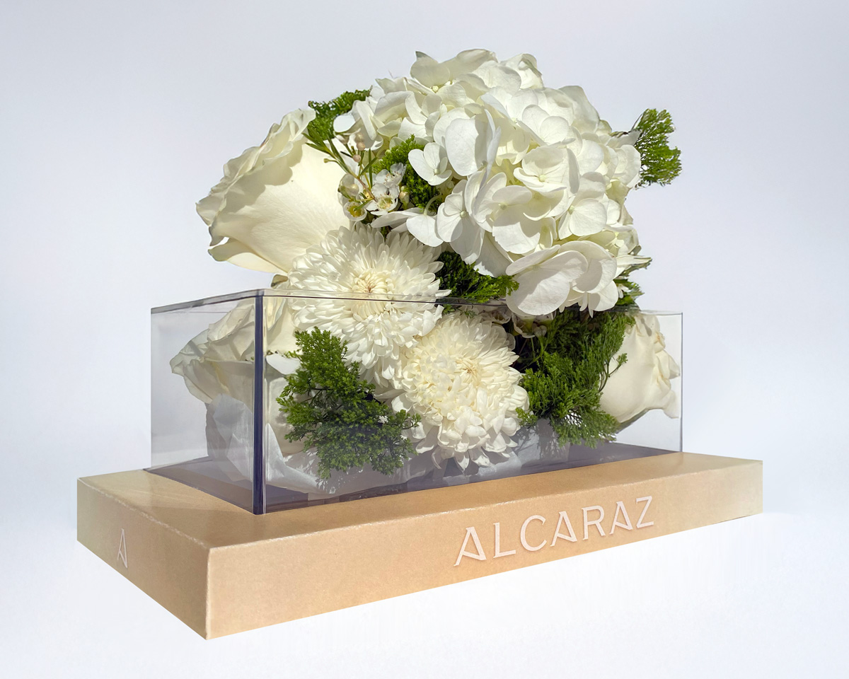 Arrangement of white flowers including roses and hydrangeas in a clear rectangular box atop a beige gradient box labeled ALCARAZ.
