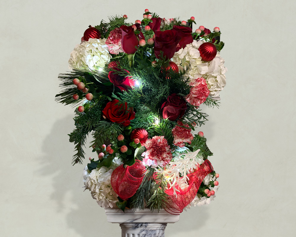 Festive Christmas floral arrangement with red roses, white hydrangeas, pink carnations, greenery, red ornaments, and lights on a marble pedestal.