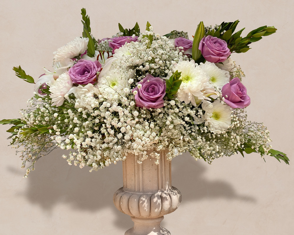 Floral arrangement of lavender roses, white chrysanthemums, baby's breath, and green foliage in a beige decorative vase.
