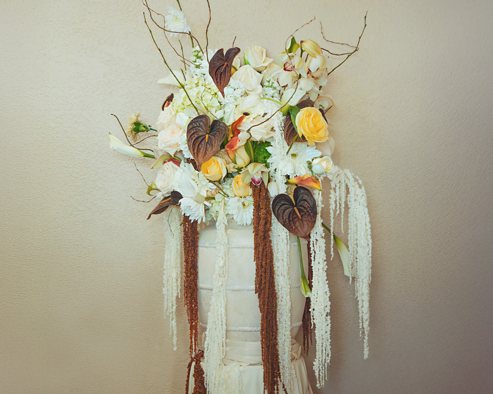 Tall floral arrangement with yellow roses, white flowers, dark brown anthuriums, and cascading white and brown hanging amaranthus in a beige vase against a plain wall.