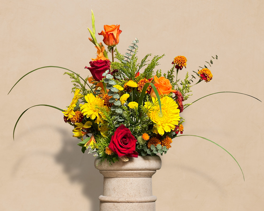 Colorful autumn floral arrangement with red and orange roses, yellow daisies, and greenery in a stone vase against a beige background.