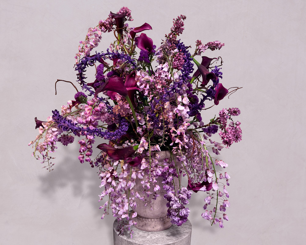Large bouquet of purple, lavender, and dark violet flowers arranged in a rustic ceramic vase on a stone pedestal against a plain light background.