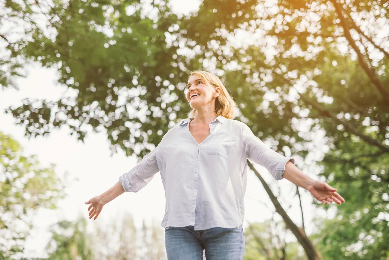 A woman smiling with her arms outstretched beneath sunlit trees, expressing a sense of peace, freedom, and renewed hope.