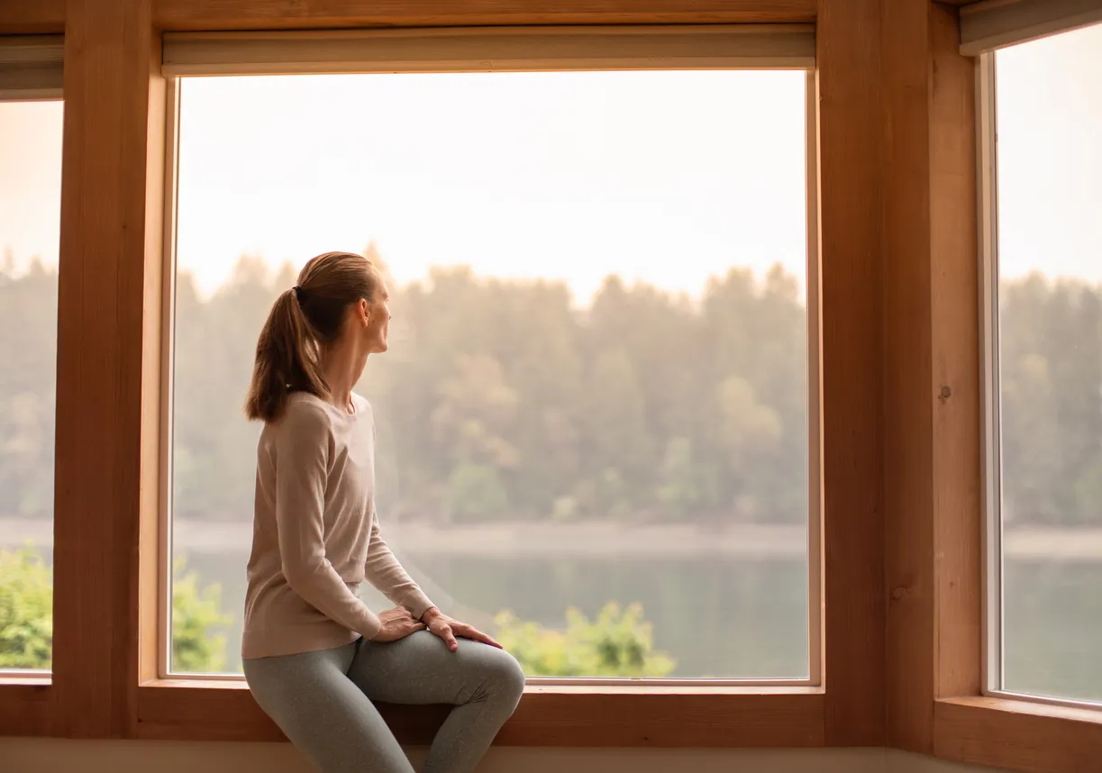 A woman sitting quietly by a large window gazing out at a peaceful lake, reflecting the stillness and hope that depression therapy can bring.