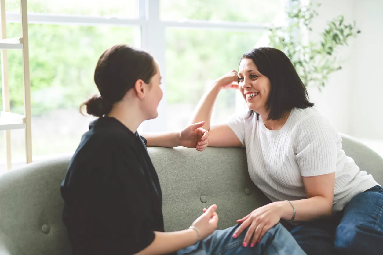 A mother and teenage daughter sharing a relaxed, smiling conversation on a couch, reflecting the stronger connection that teen therapy can build.