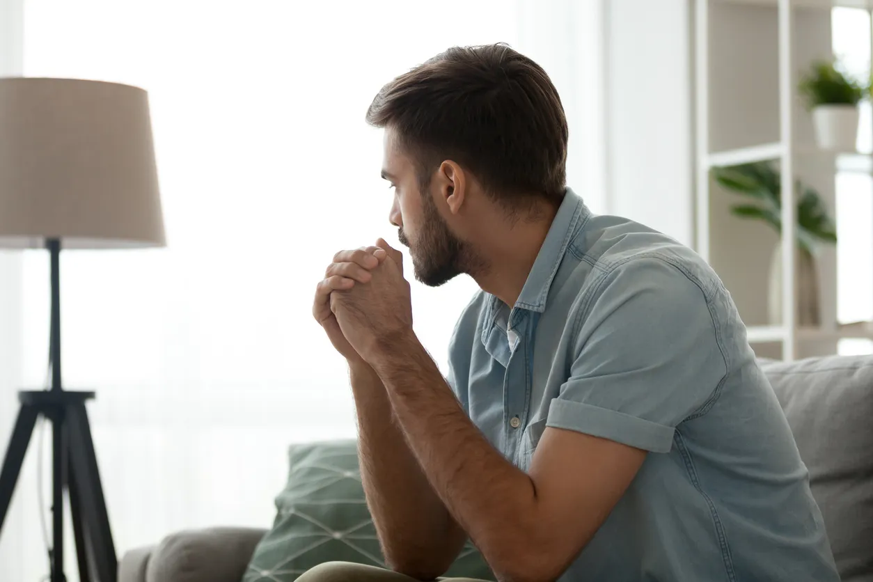 A man sitting on a couch with his hands clasped, deep in thought, representing the self-reflection that anger management therapy encourages.