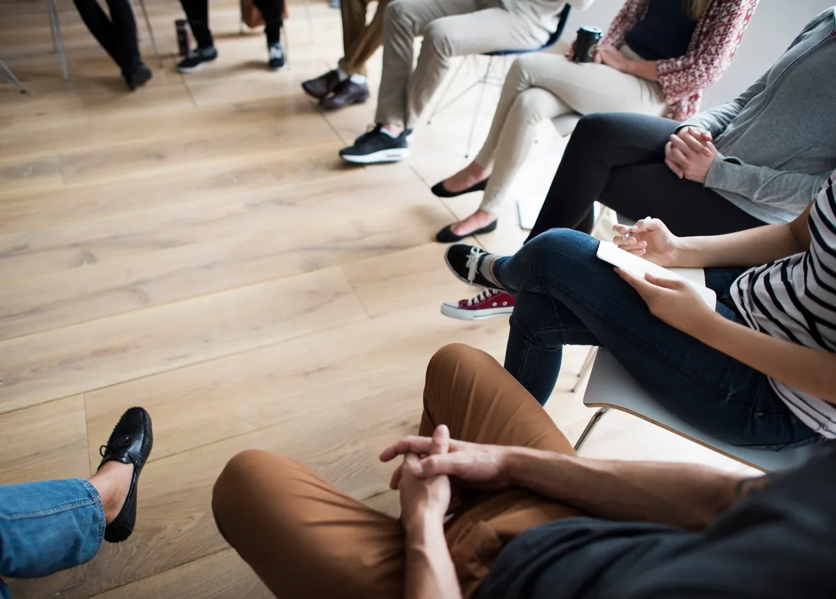 A group of people seated in a circle during a group therapy session, with hands resting naturally as they listen and share in a supportive setting.