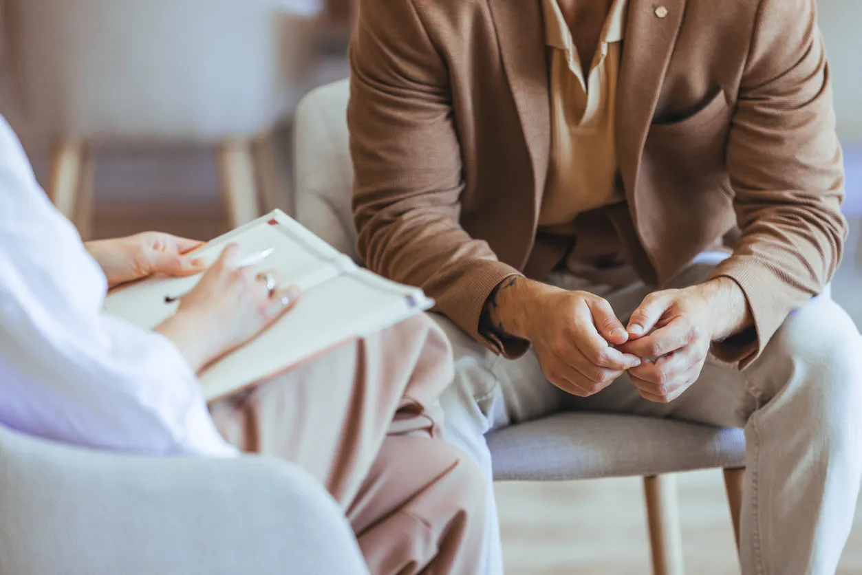 A patient sits with hands gently clasped while a provider holds a clipboard during a psychiatric evaluation in a calm, professional setting.