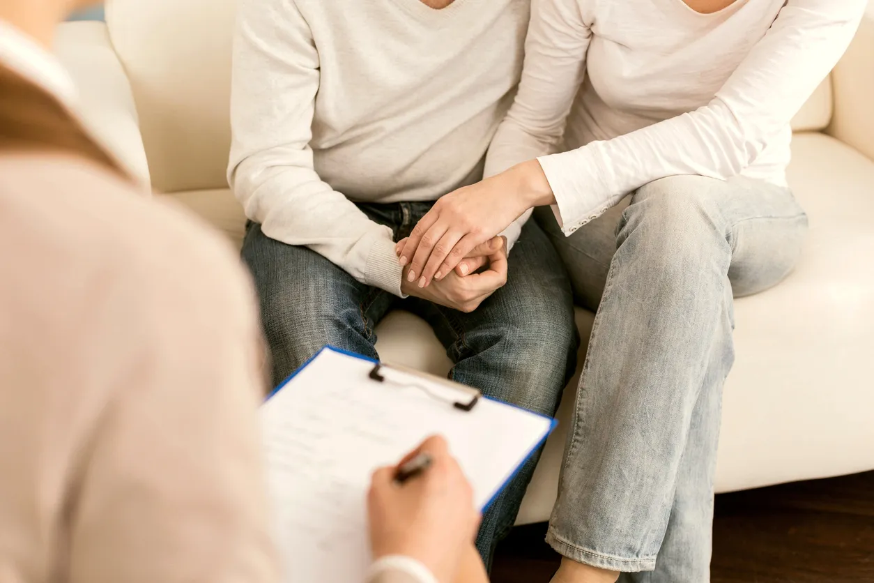 An engaged couple holds hands on a couch across from a counselor with a clipboard, working together through premarital counseling to build a strong foundation before their wedding.
