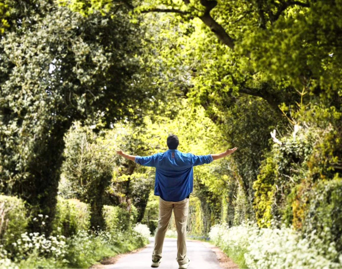 Person standing on a tree-lined path with arms outstretched, surrounded by lush green foliage.