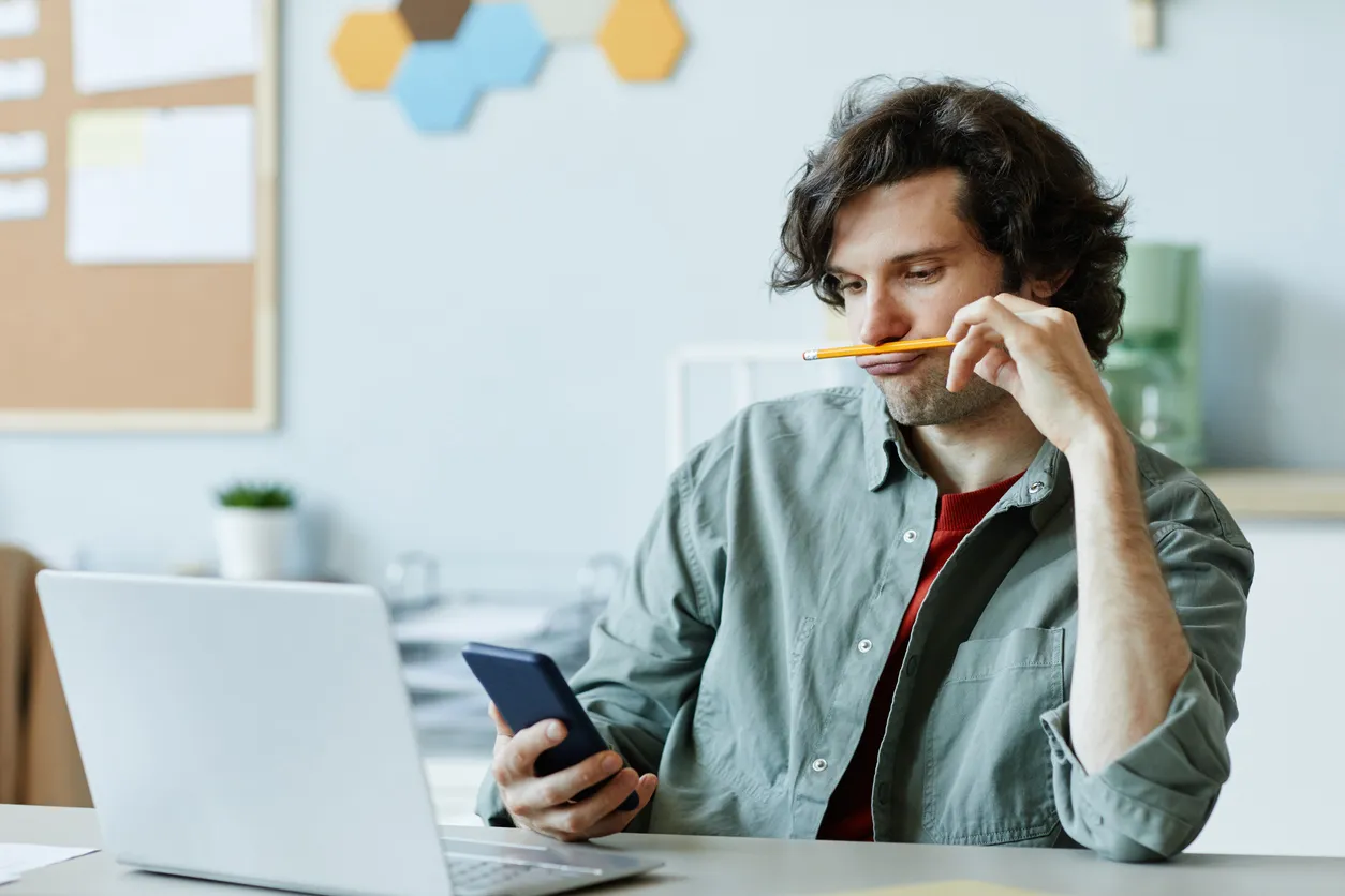 A man holds a pencil to his face while looking at his phone at a desk with a laptop open, illustrating the difficulty with focus that ADHD treatment can help manage.
