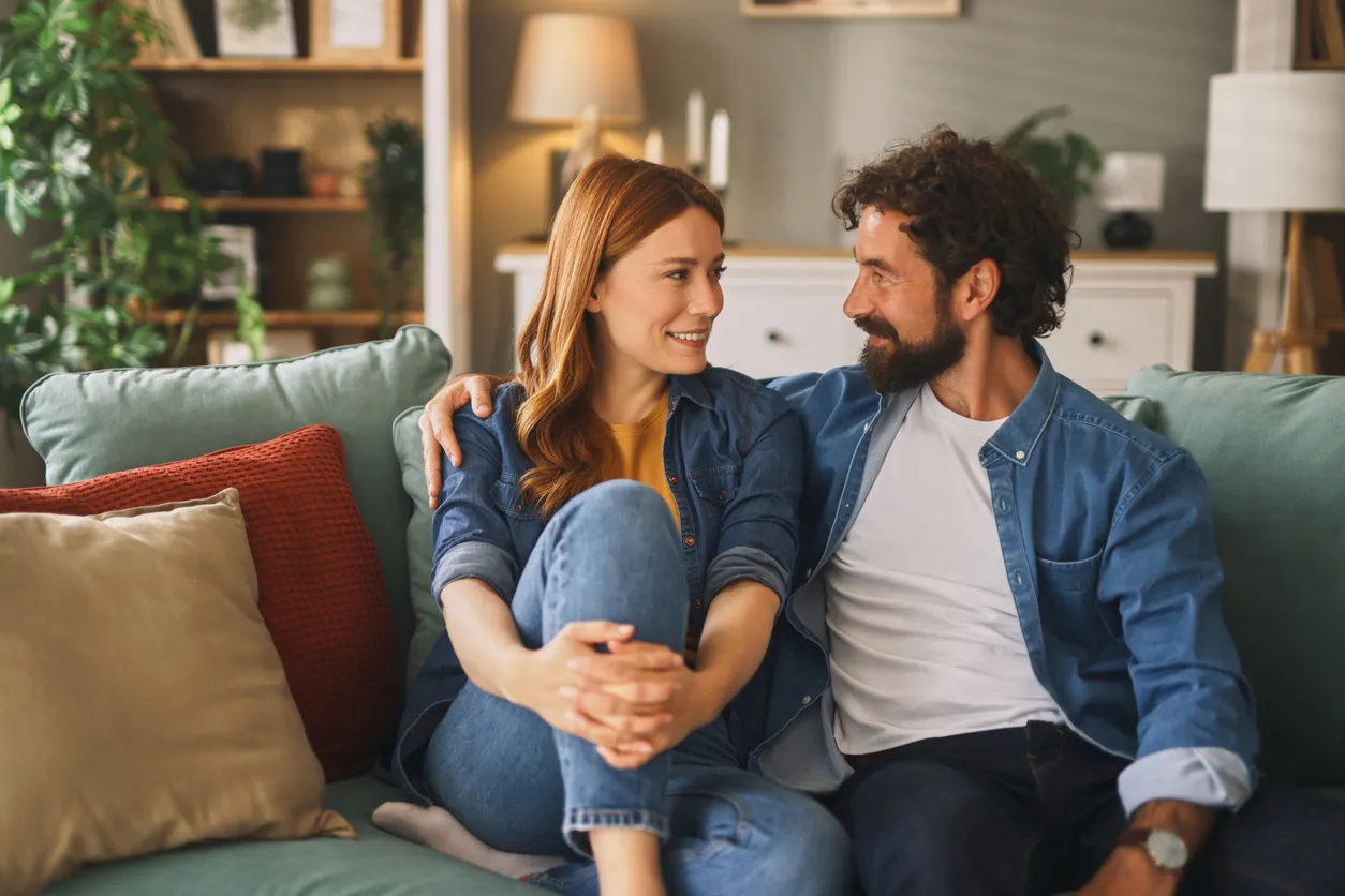 A smiling couple sitting close together on their living room couch, enjoying the kind of open, connected conversation that couples workshops help cultivate.