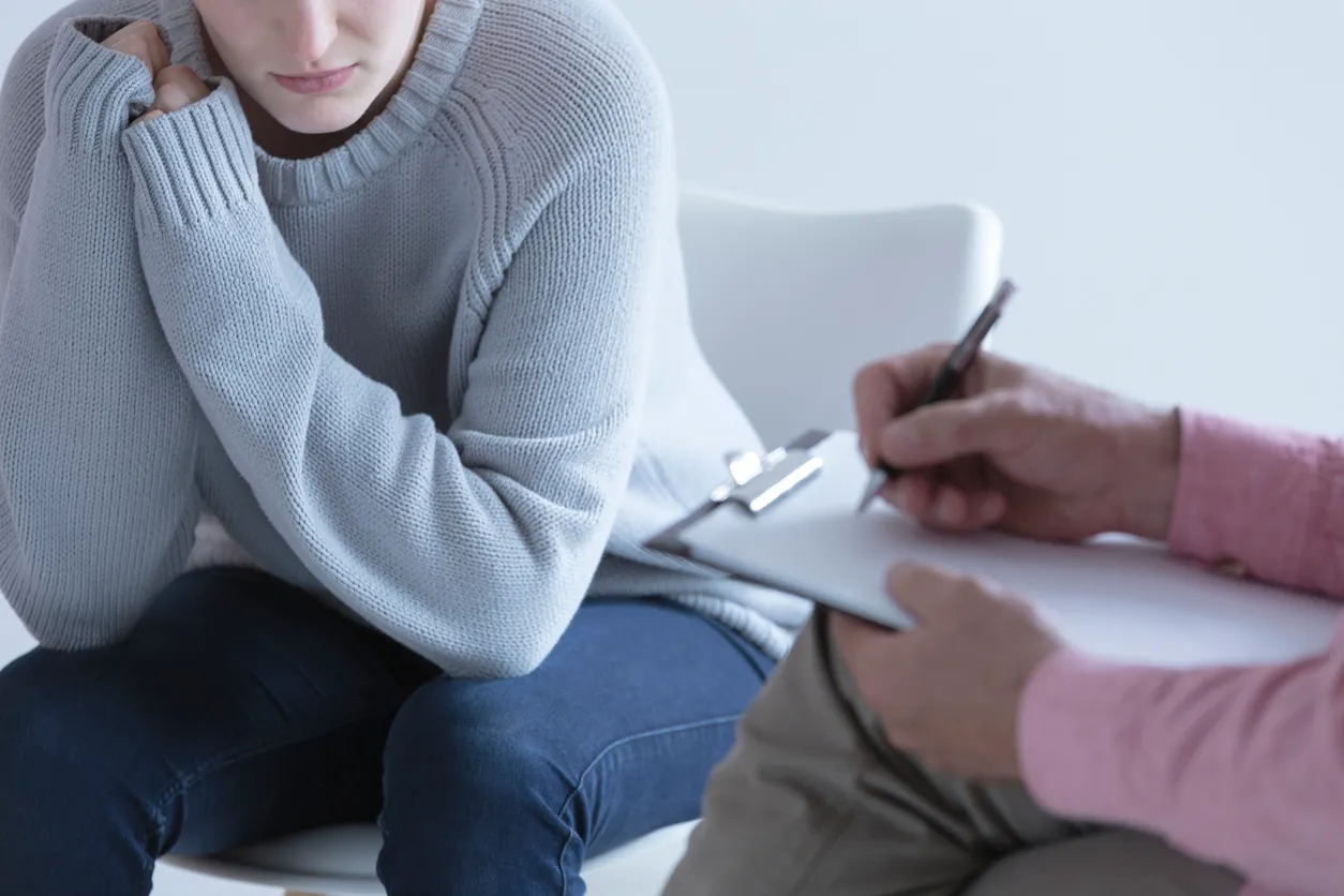 A patient sits with arms drawn close during an anxiety psychiatry session while a provider takes notes nearby, capturing the vulnerability and care involved in seeking help.
