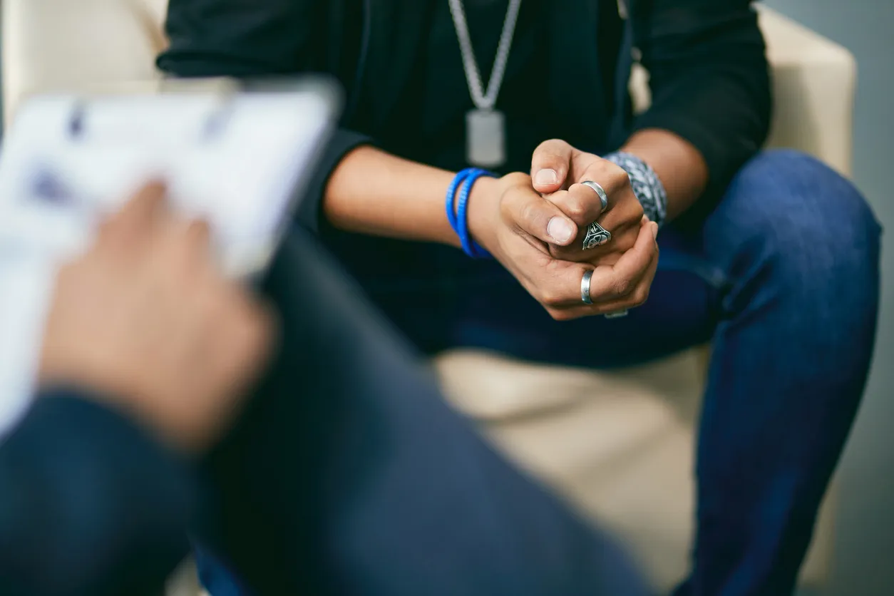 A young person sits with hands clasped during a bipolar disorder psychiatry session while a provider takes notes, reflecting the trust and openness that treatment requires.