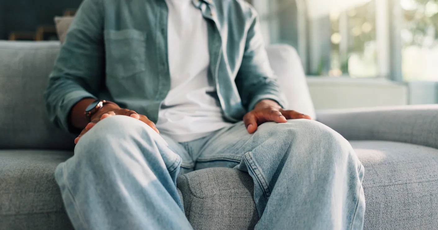 A man sitting on a gray couch with his hands resting on his knees in a sunlit living room, ready to take the first step toward schizophrenia psychiatry care.
