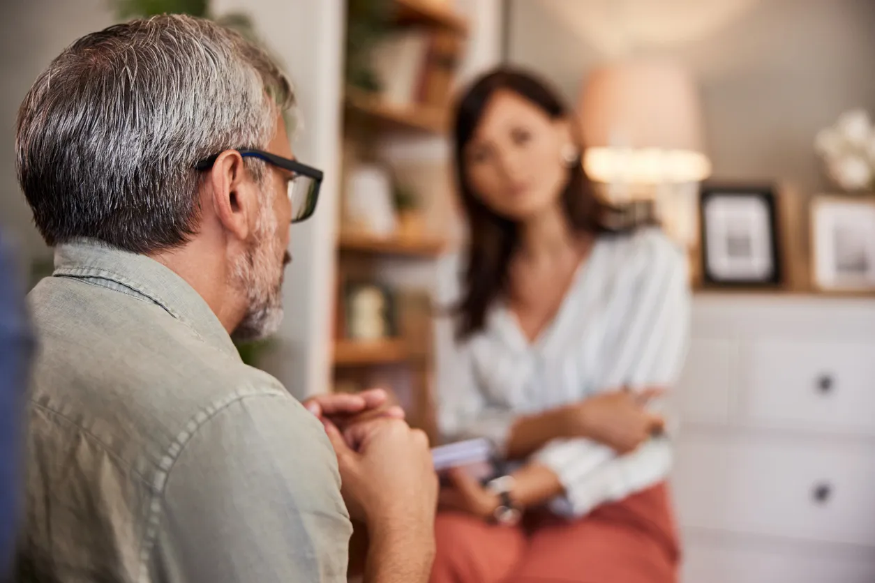 A man with glasses listens attentively to his therapist during a counseling session in a warm, comfortable office.