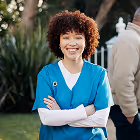 Smiling woman with curly hair wearing teal medical scrubs outdoors with arms crossed.