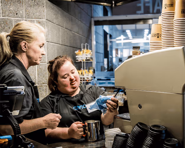 Two women, one with a disability, working together making coffee at a cafe using an espresso machine.