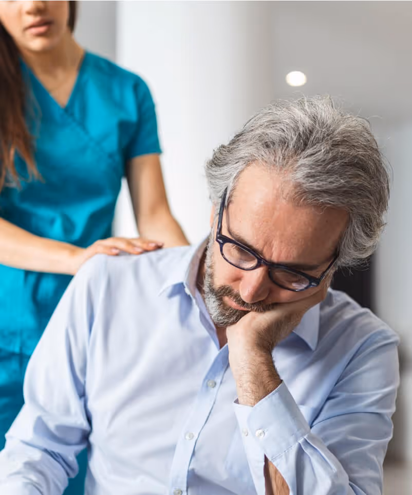 Healthcare worker comforting a stressed middle-aged man with glasses by placing a hand on his shoulder.