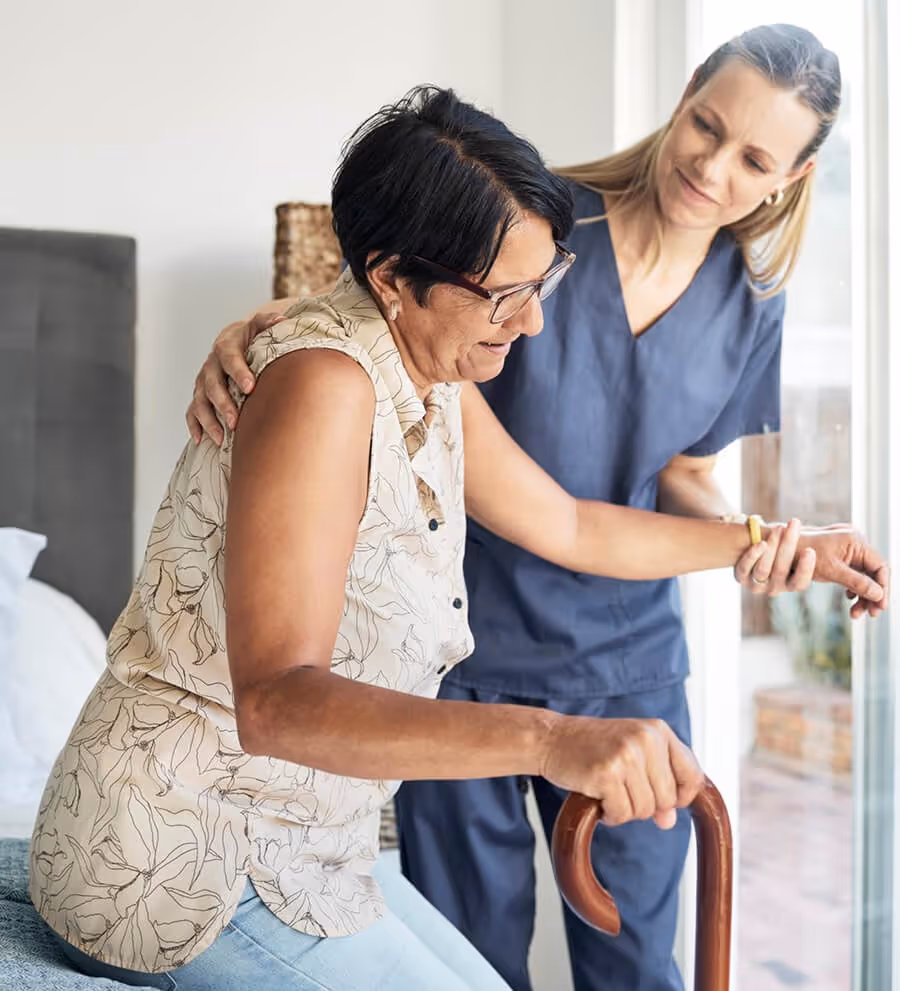 Caregiver in blue scrubs assisting elderly woman holding a wooden cane as she stands by a window.