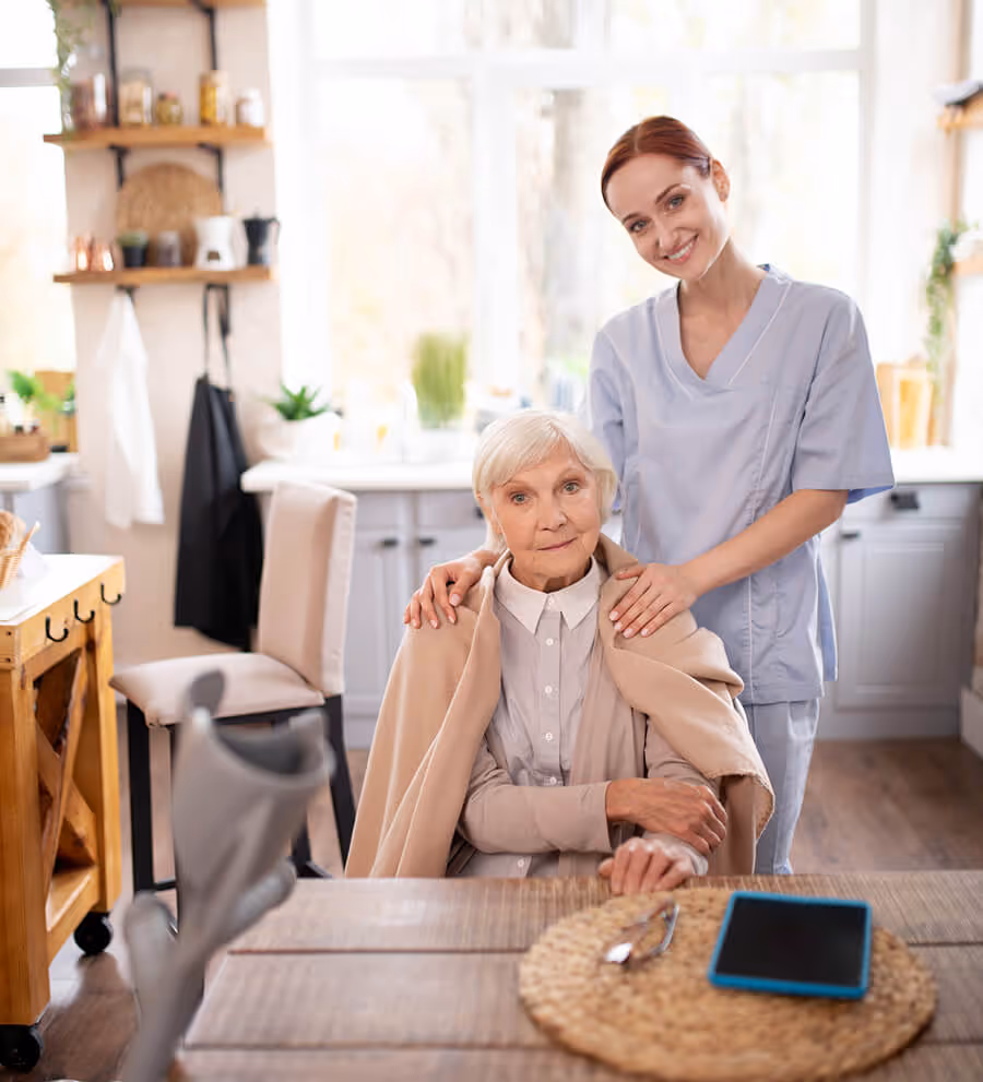 Smiling female caregiver standing behind an elderly woman wrapped in a beige blanket, in a bright, cozy kitchen.
