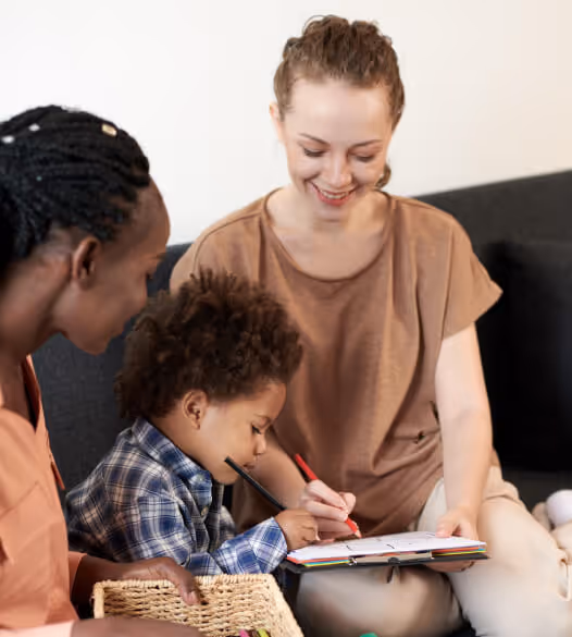 Two women and a young boy sitting together on a couch, the boy drawing with colored pencils while one woman smiles and the other watches.