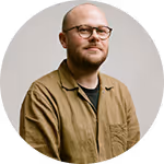 Bald man with glasses and a beard wearing a brown shirt against a plain background.
