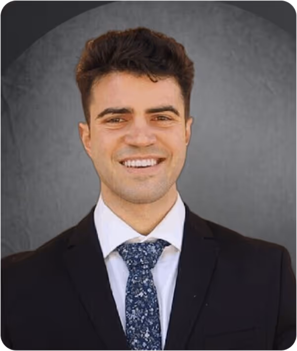 Smiling young man wearing a dark suit, white shirt, and blue floral tie against a gray background.