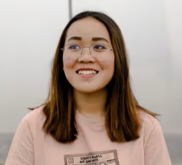 Smiling young woman with glasses and shoulder-length brown hair wearing a light pink t-shirt.