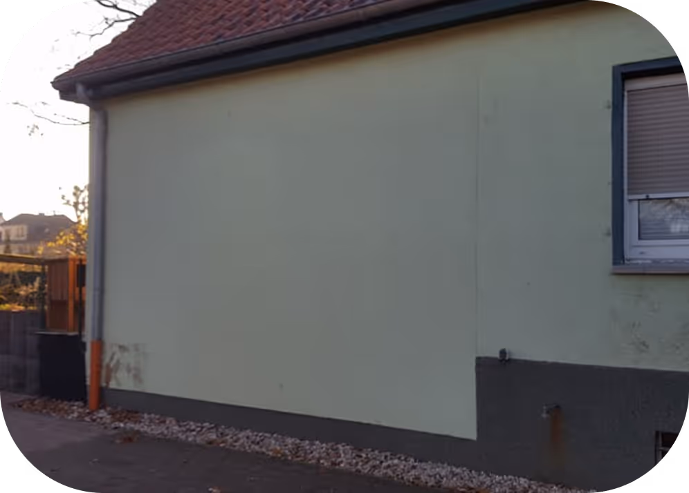 Außenwand eines Hauses mit grünlicher Fassade, grauem Sockel, geschlossenem Fenster mit Rollladen und Dach mit roten Ziegeln.