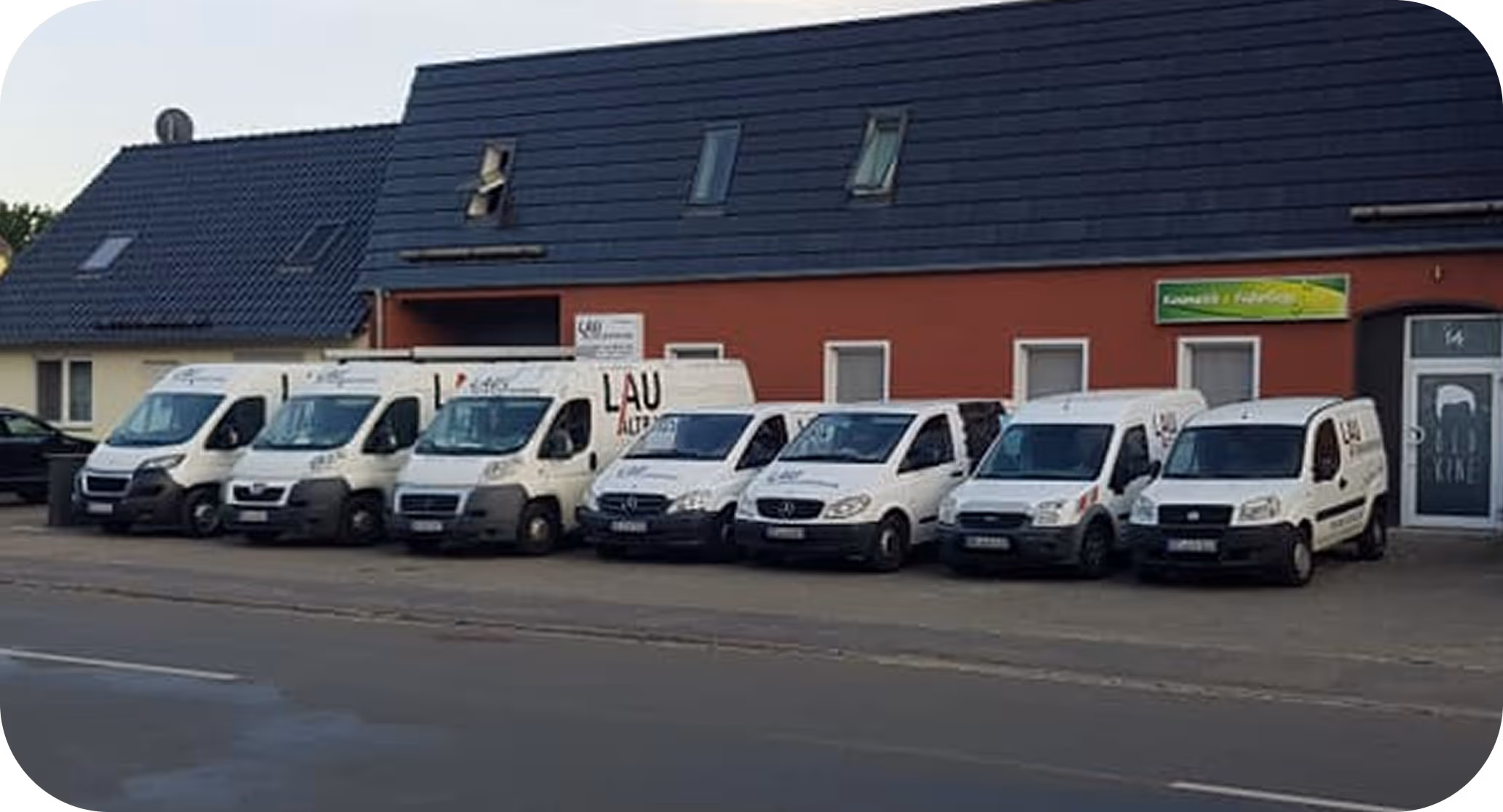 Sieben weiße Lieferwagen stehen geparkt vor einem Gebäude mit roter Fassade und dunklem Dach.