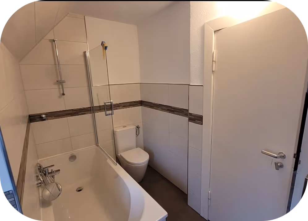 Kleine weiße Badezimmer mit Badewanne, Glasdusche und Toilette, weiß gekachelte Wände mit braunem Streifen.