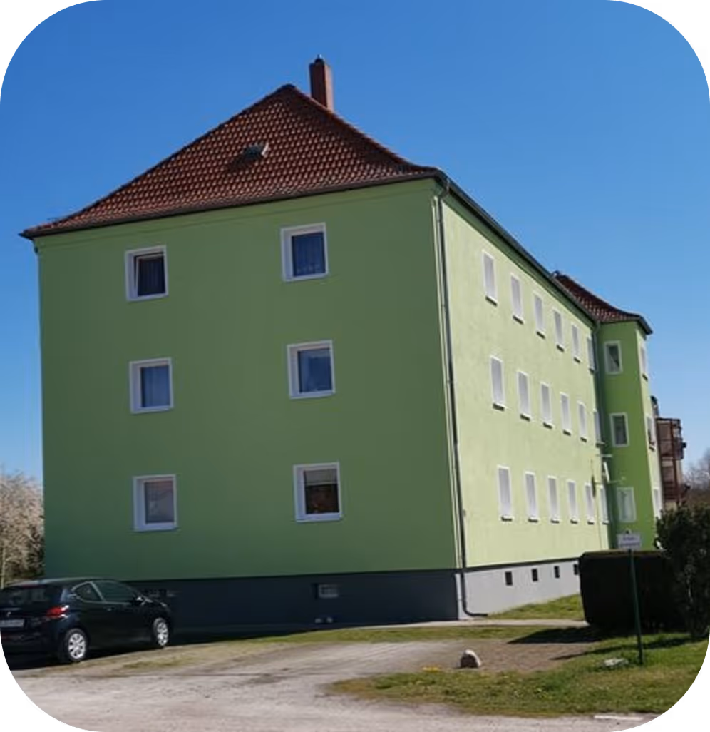Dreistöckiges grünes Wohngebäude mit rotem Ziegeldach und geparktem schwarzem Auto davor unter blauem Himmel.