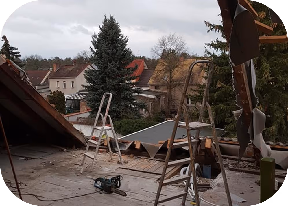 Innenraum eines teilweise abgedeckten Dachs mit zwei Leitern und einer Kettensäge, Blick auf Häuser und Bäume im Hintergrund.