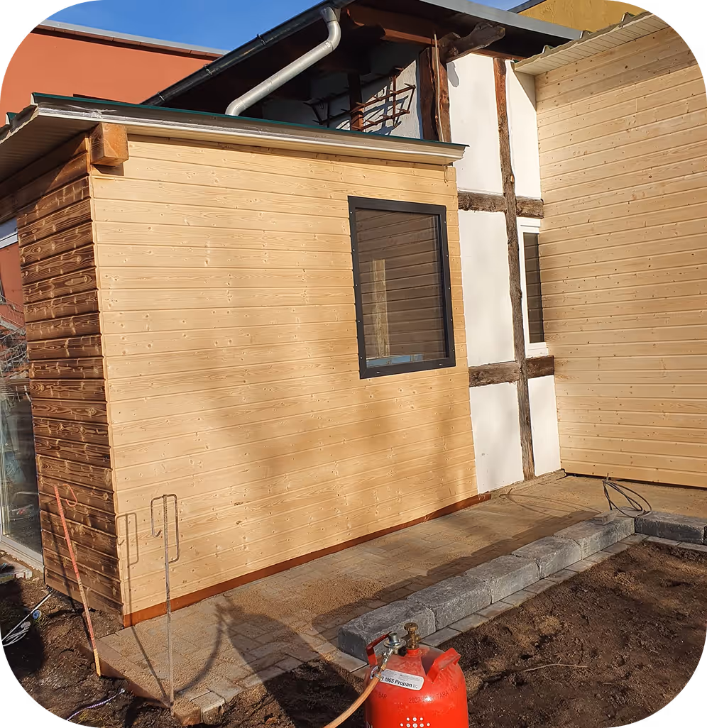 Holzverkleideter Anbau an einem Fachwerkhaus mit Fenster und rotem Propangasflasche im Vordergrund.