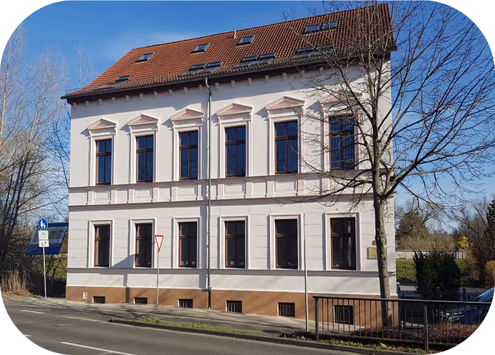 Historisches zweistöckiges Gebäude mit weißen Wänden, braunen Fensterrahmen und rotem Ziegeldach an einer Straße bei sonnigem Wetter.