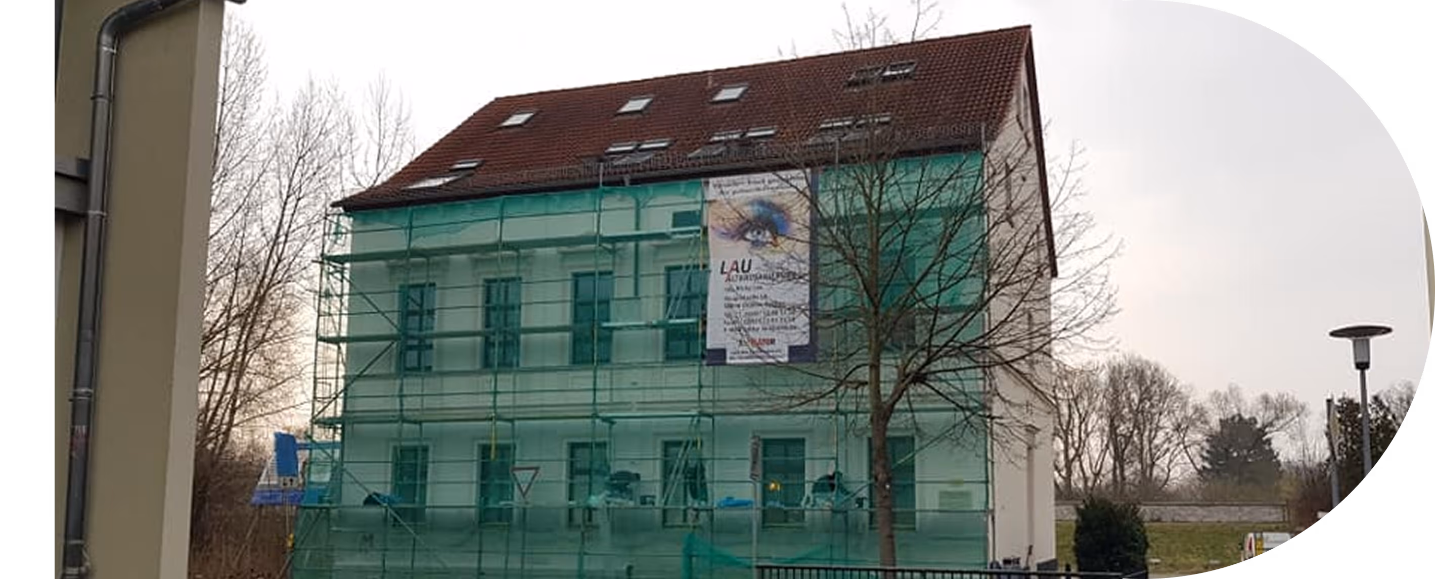 Gebäude mit grünem Gerüstnetz und einem Banner mit Augenwerbung an der Fassade, umgeben von kahlen Bäumen und einer Laterne an einem bewölkten Tag.