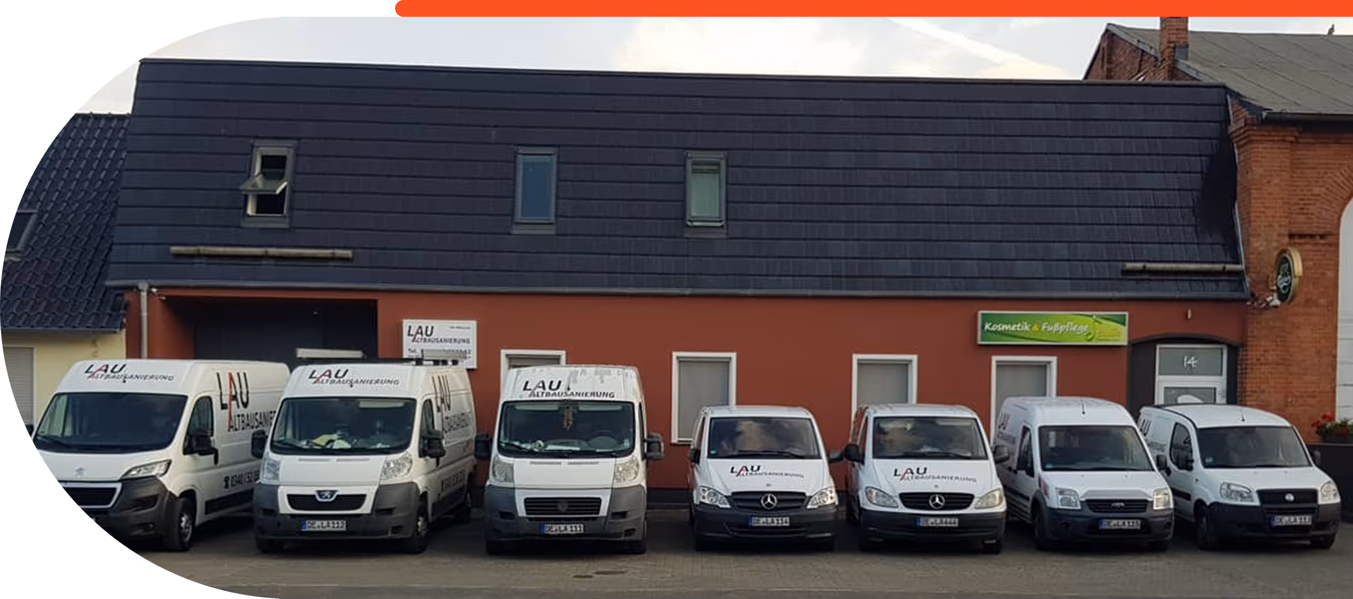 Siebene weiße Firmenfahrzeuge mit dem Logo LAU Altbausanierung parken vor einem roten Gebäude mit dunklem Dach.