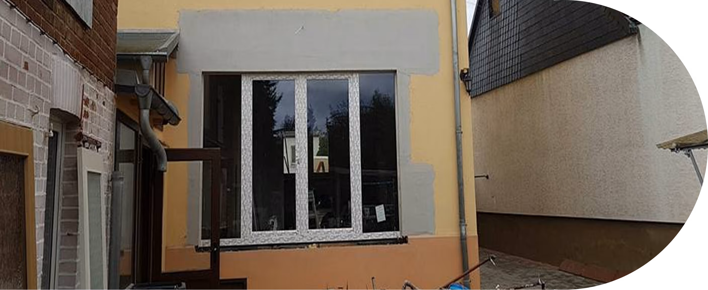 Hausfassade mit neu installierten weißen Kunststofffenstern, umgeben von gelber und oranger Putzfassade mit grauen Ausbesserungsstellen.