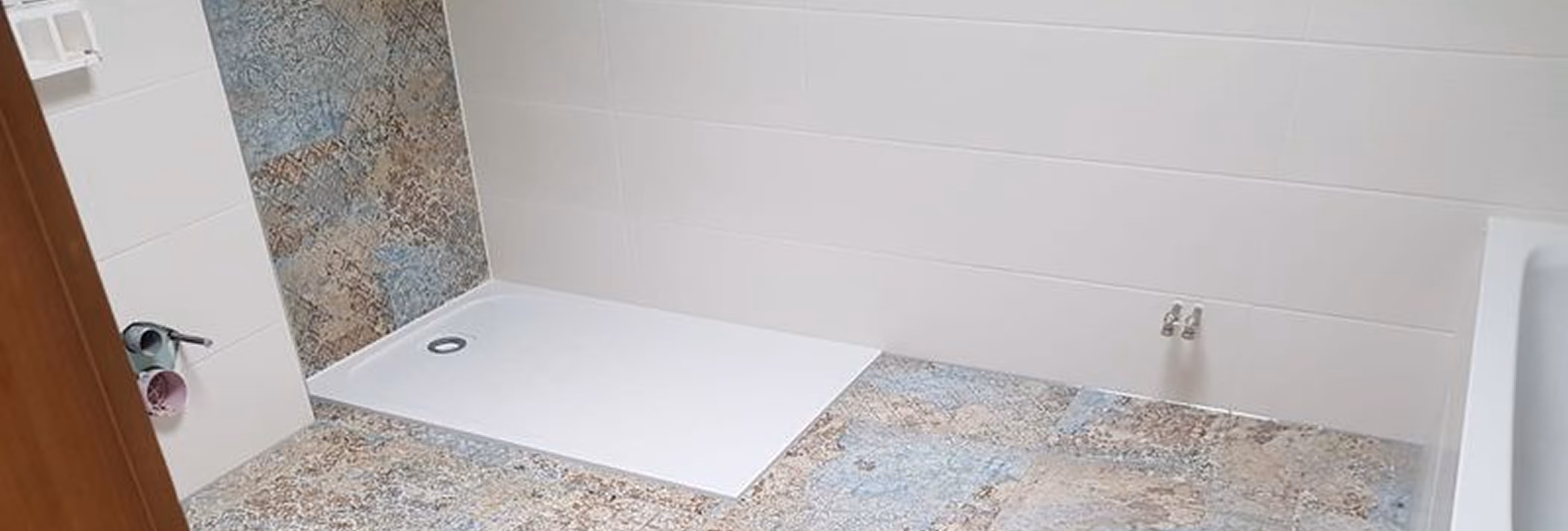 Badezimmer mit einer weißen Duschwanne, weißen Wandfliesen und gemusterten Bodenfliesen in Blau- und Brauntönen.