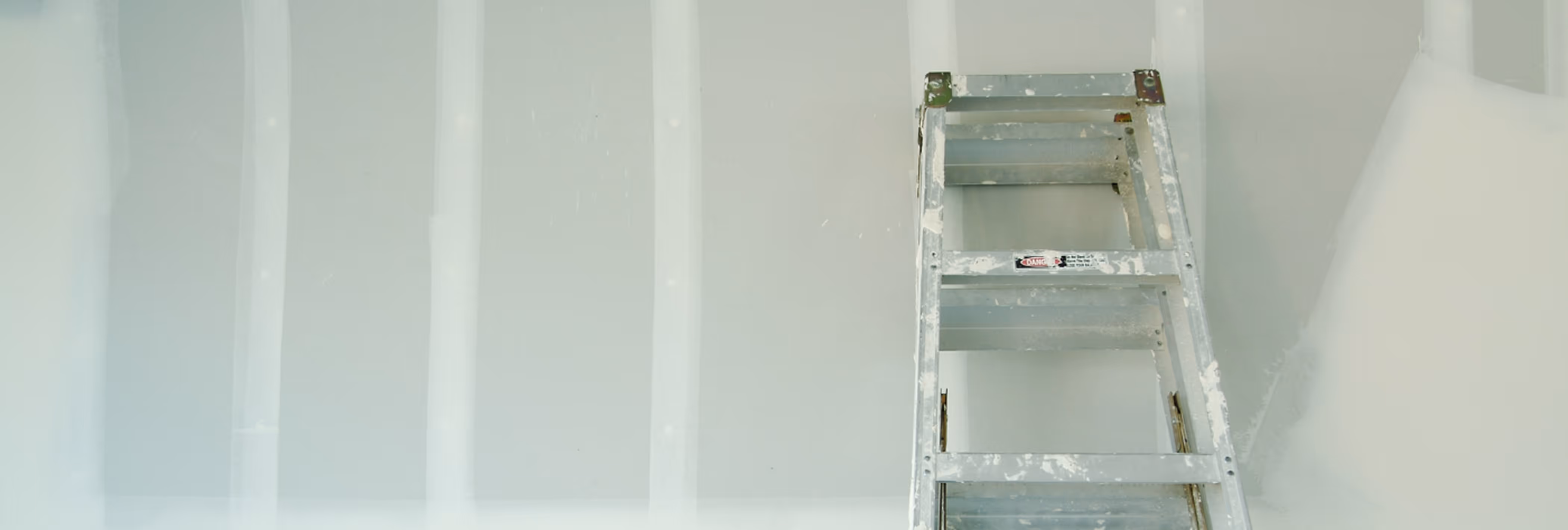 Leiter mit weißen Farbflecken vor einer hellgrauen Wand im Bauzustand.